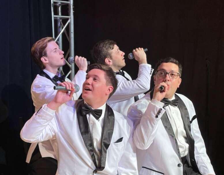 A group of four young men in matching white suits, all singing